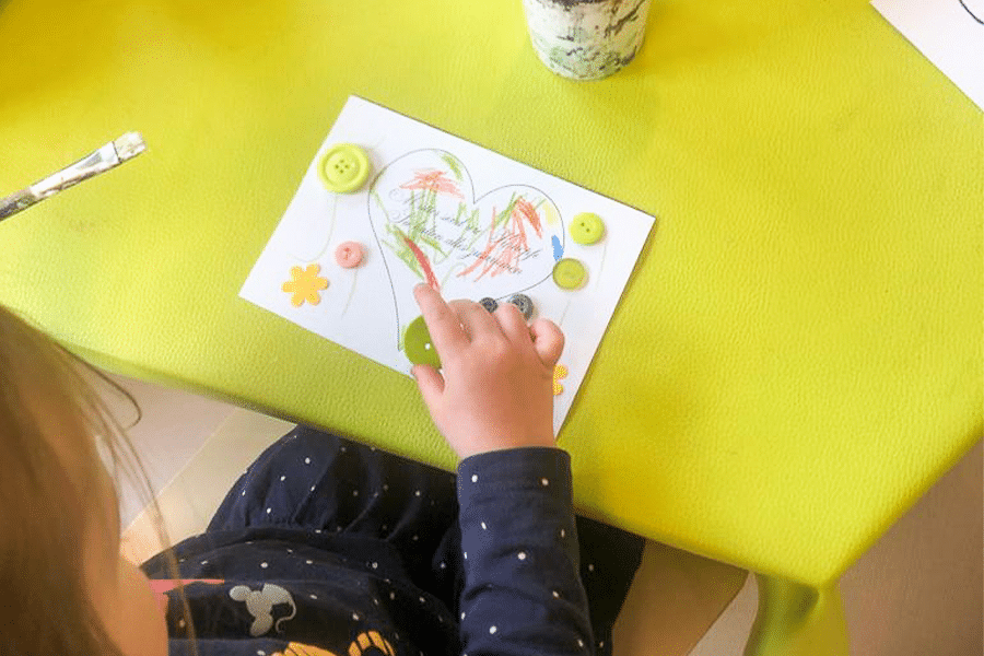 Kind macht Muttertagsgeschenk auf Tisch mit Knöpfen und Bastelutensilien